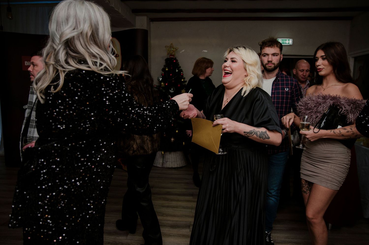 Two people in sparkly evening wear laugh and dance together at an indoor party while others watch in the background.
