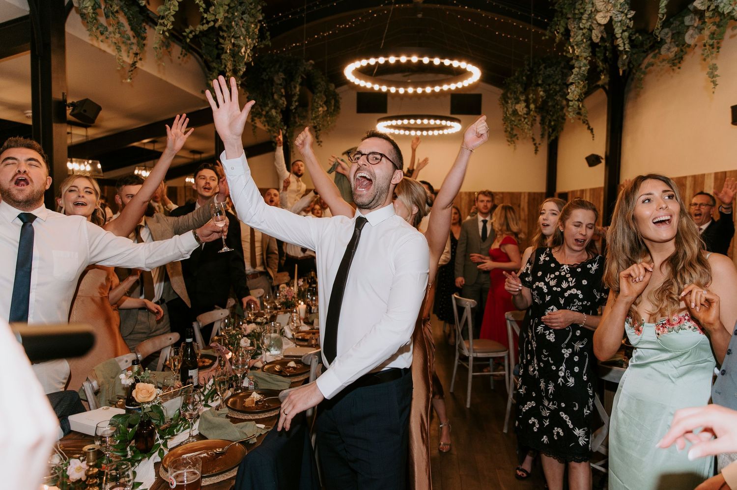 Wedding guests dance and celebrate enthusiastically on a dance floor under string lights at an indoor reception venue.
