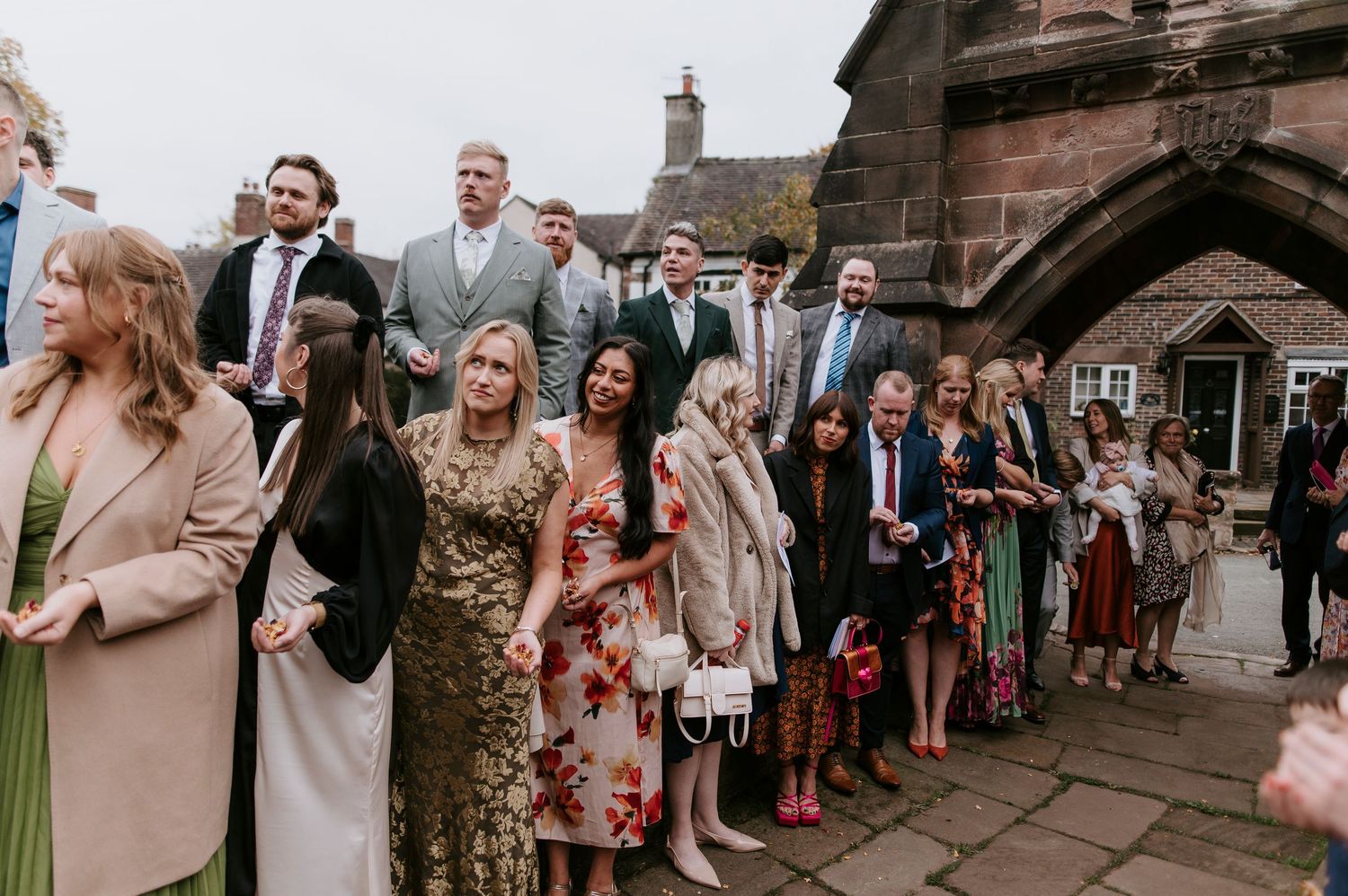 Wedding party and guests line up along stone church walls during outdoor celebration.
