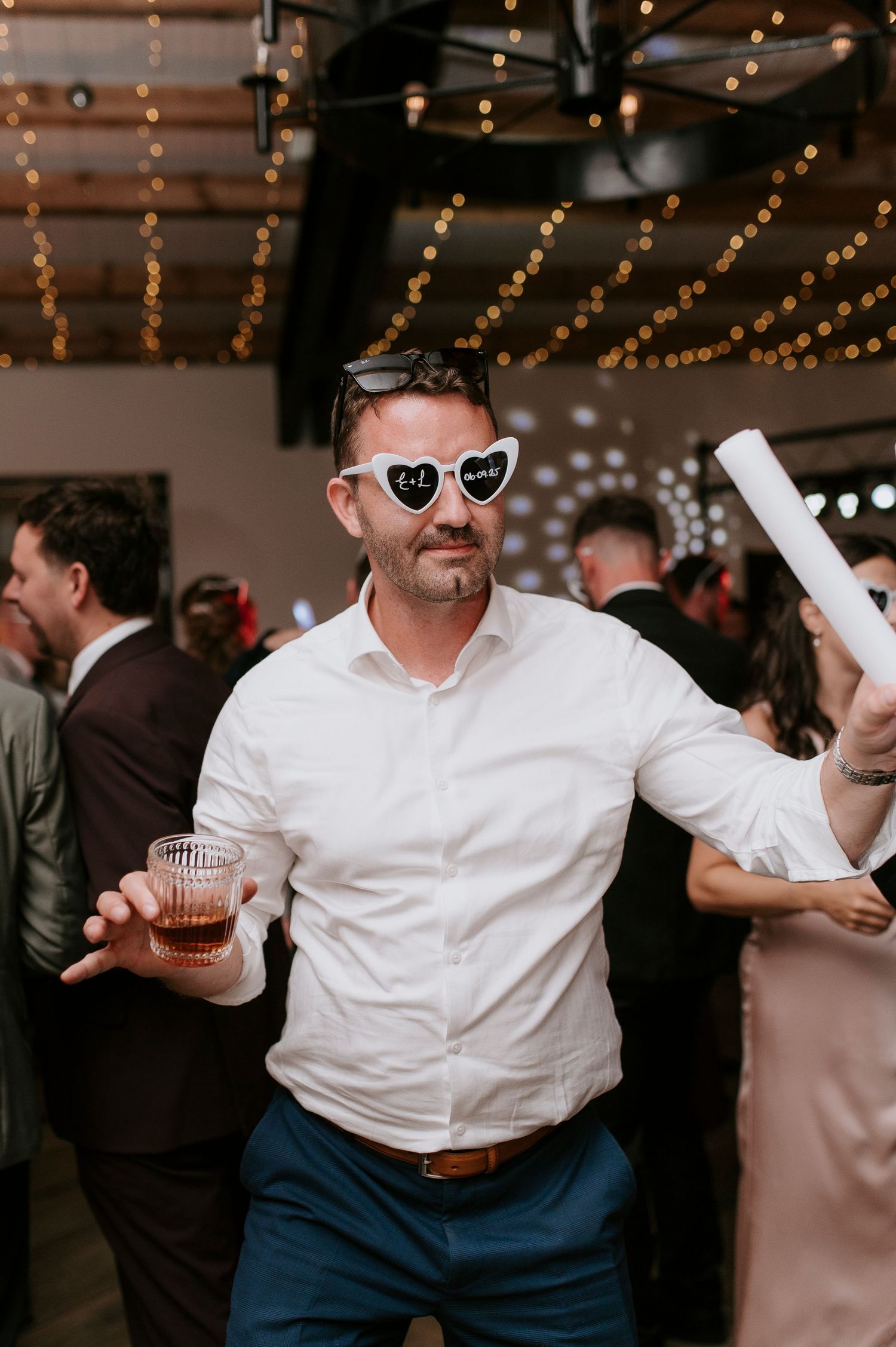 A person in a white button-down shirt and sunglasses dances energetically at an indoor event with string lights overhead.