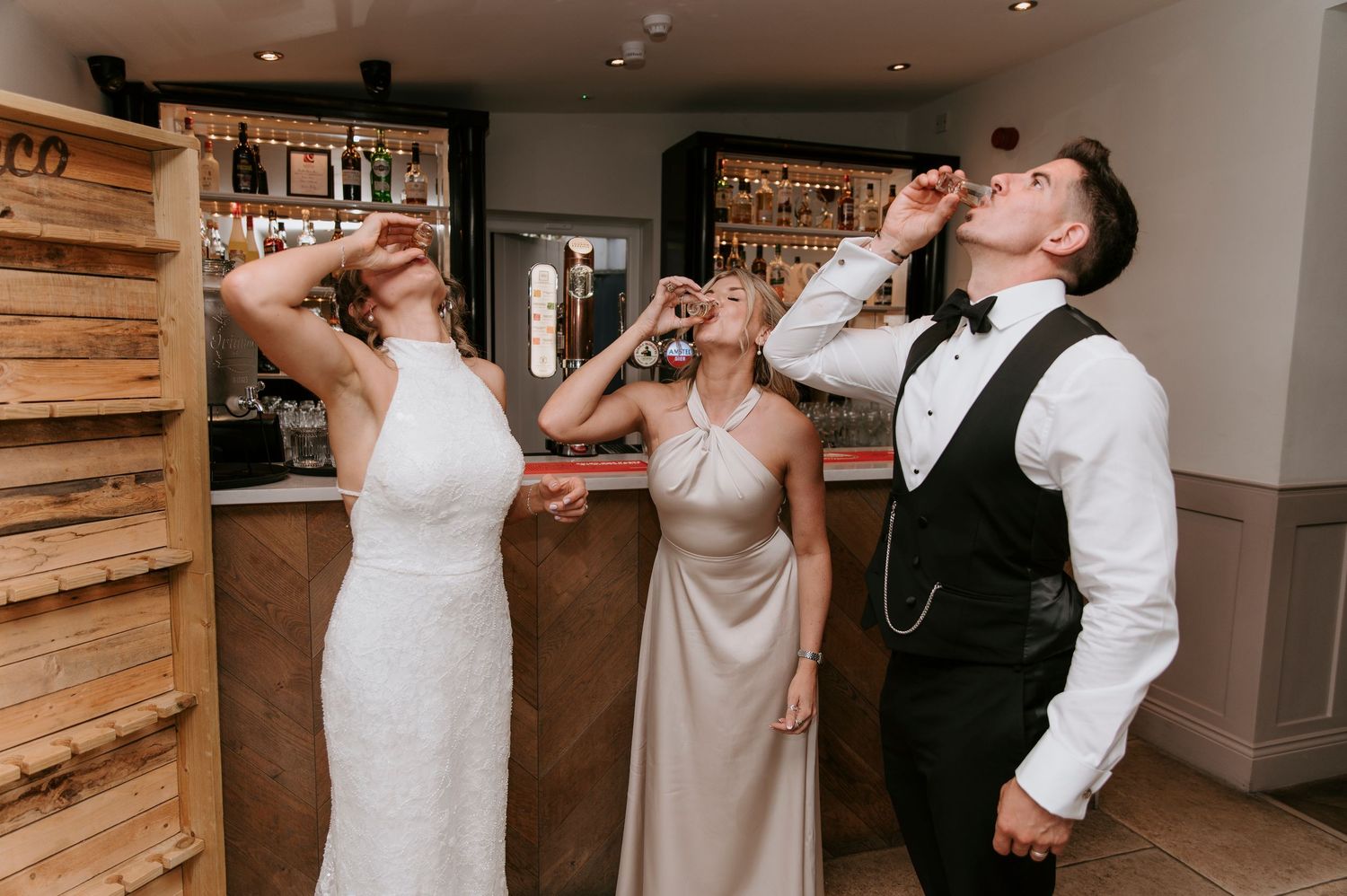 Wedding party takes celebratory shots together at reception venue with rustic wooden wall decor.