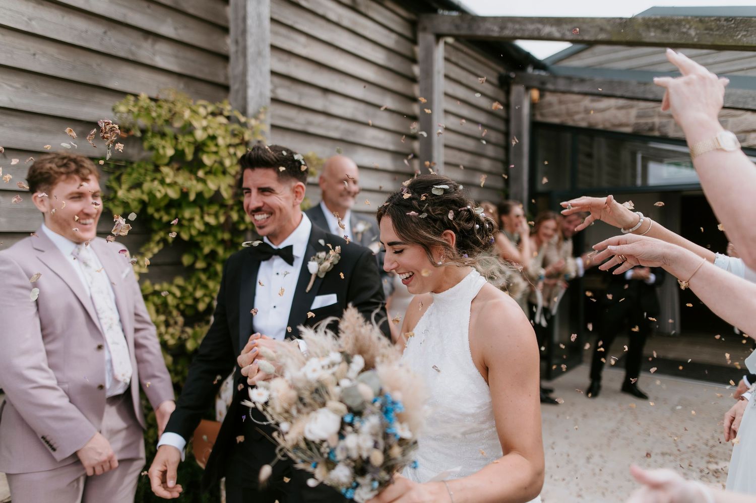 Wedding party celebrates outdoors with confetti toss as newlyweds share a joyful moment in front of rustic building.