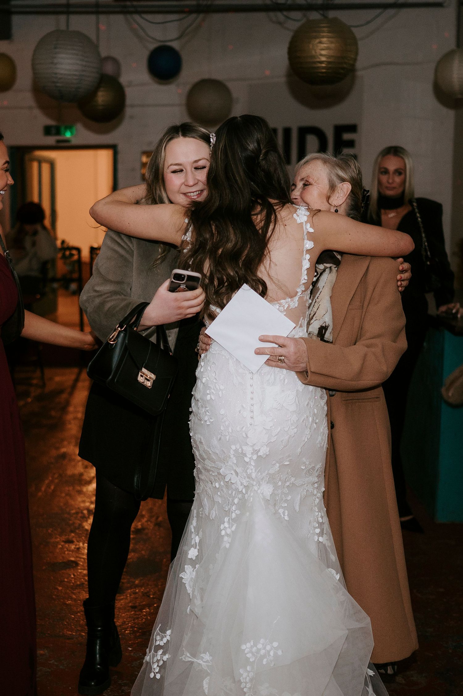 A bride in a lace wedding gown receives warm hugs from guests at her wedding reception.