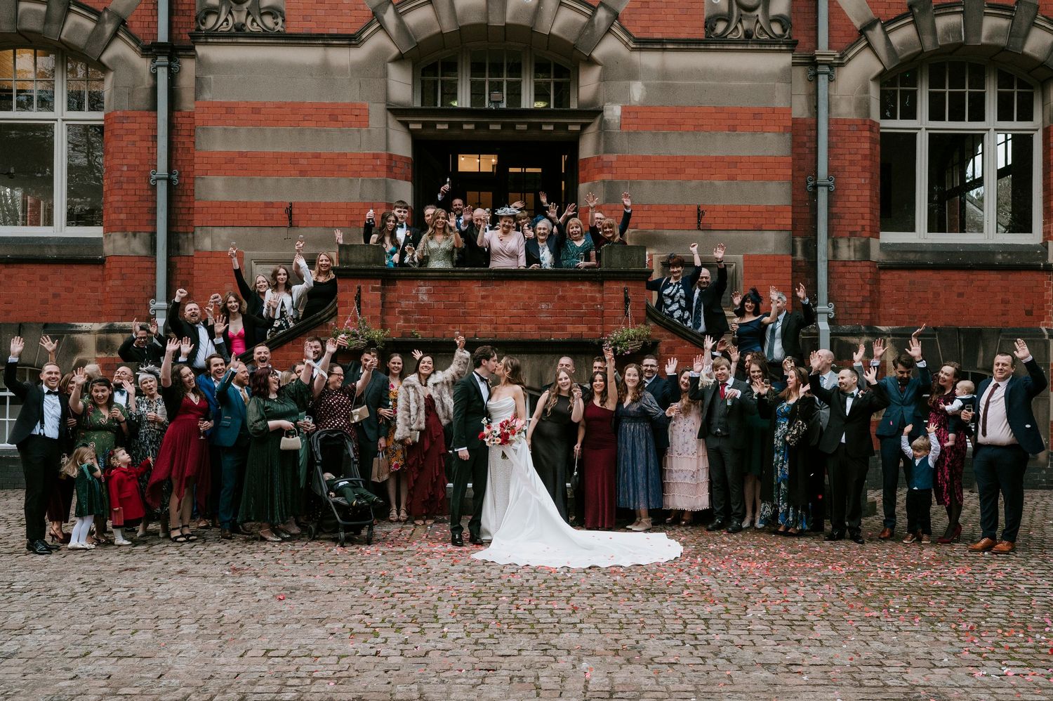 Large wedding party poses for group photo on steps of historic red brick building with scattered rose petals.