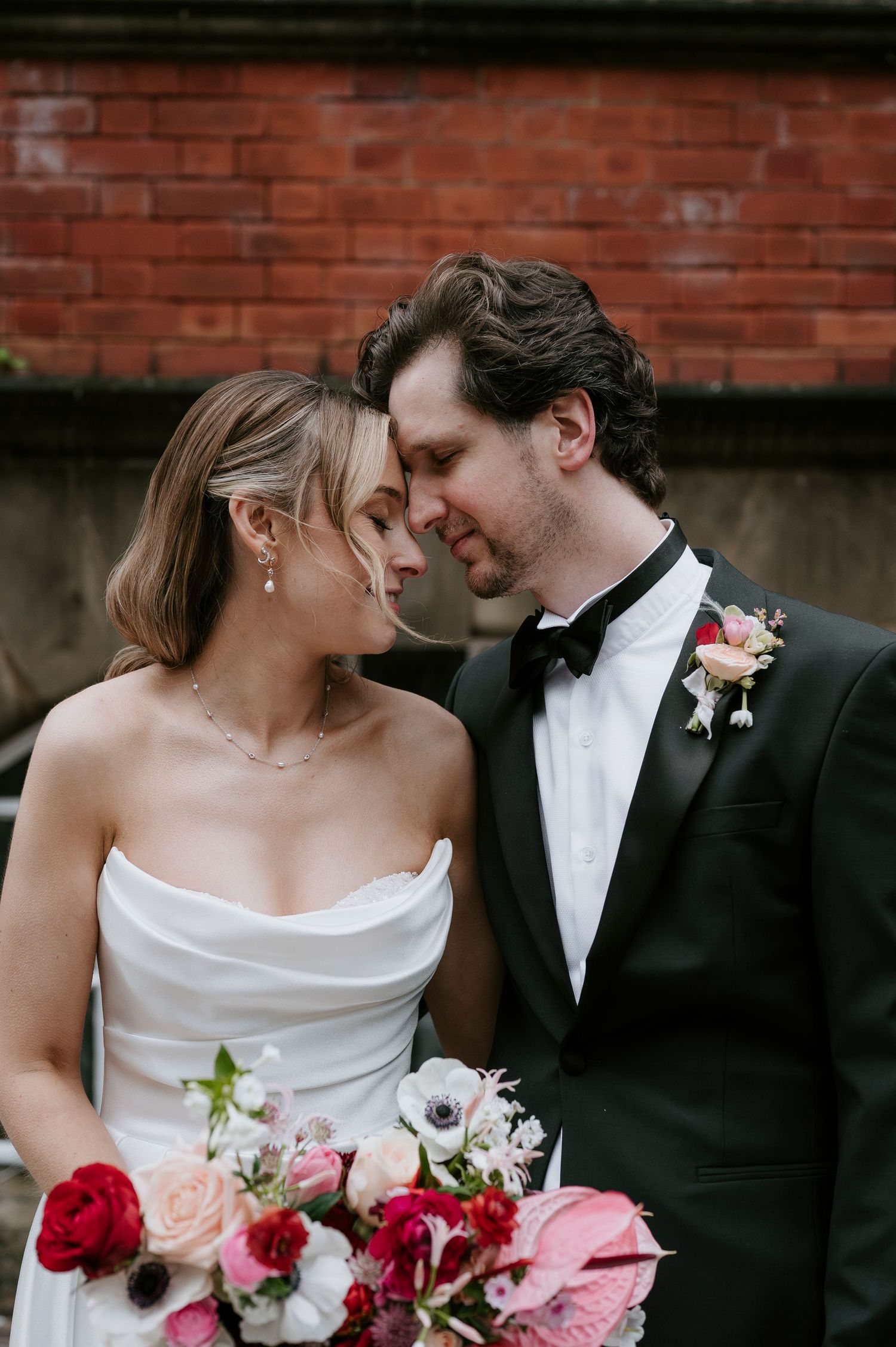 Romantic wedding couple sharing intimate moments with pink and red floral bouquet against brick wall backdrop.