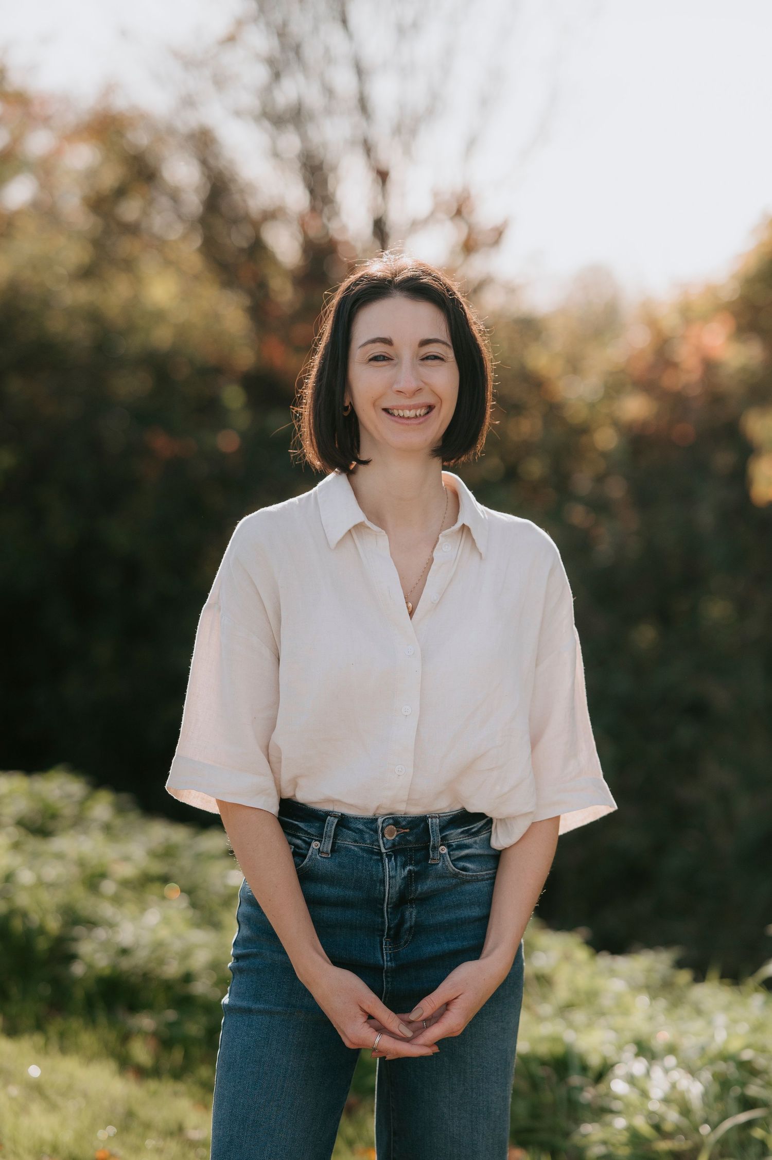 A person wearing a white button-up shirt and blue jeans poses outdoors in natural sunlight against a green background.