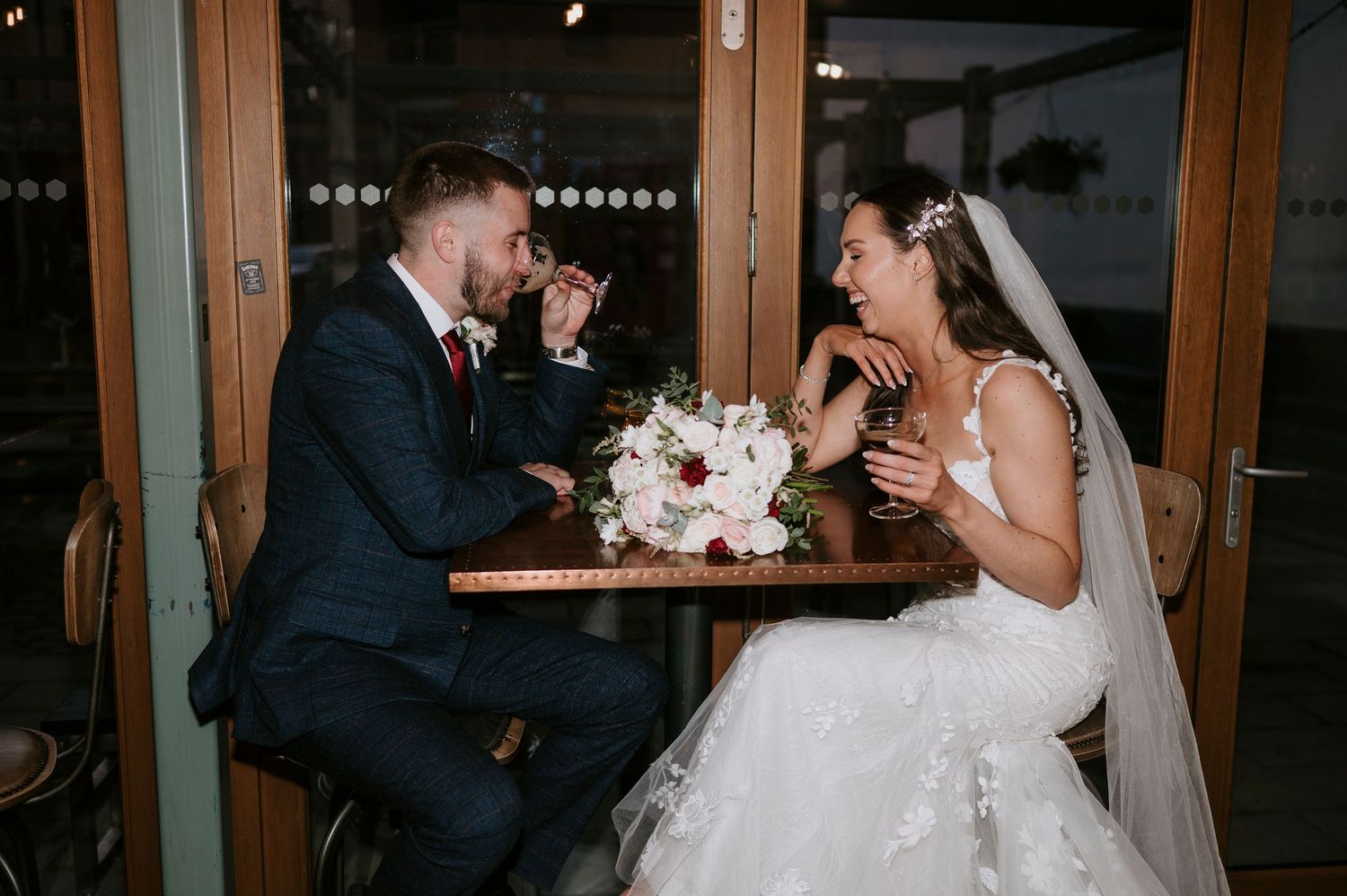 Bride and groom laugh together at a table with a floral bouquet during their wedding reception.