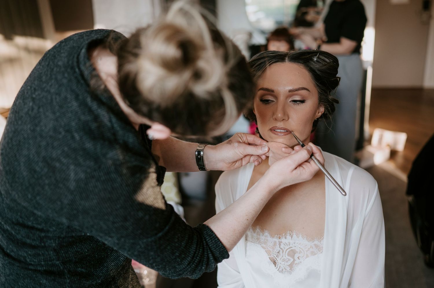 A makeup artist applies lipstick to a bride in a white lace robe during bridal preparations.