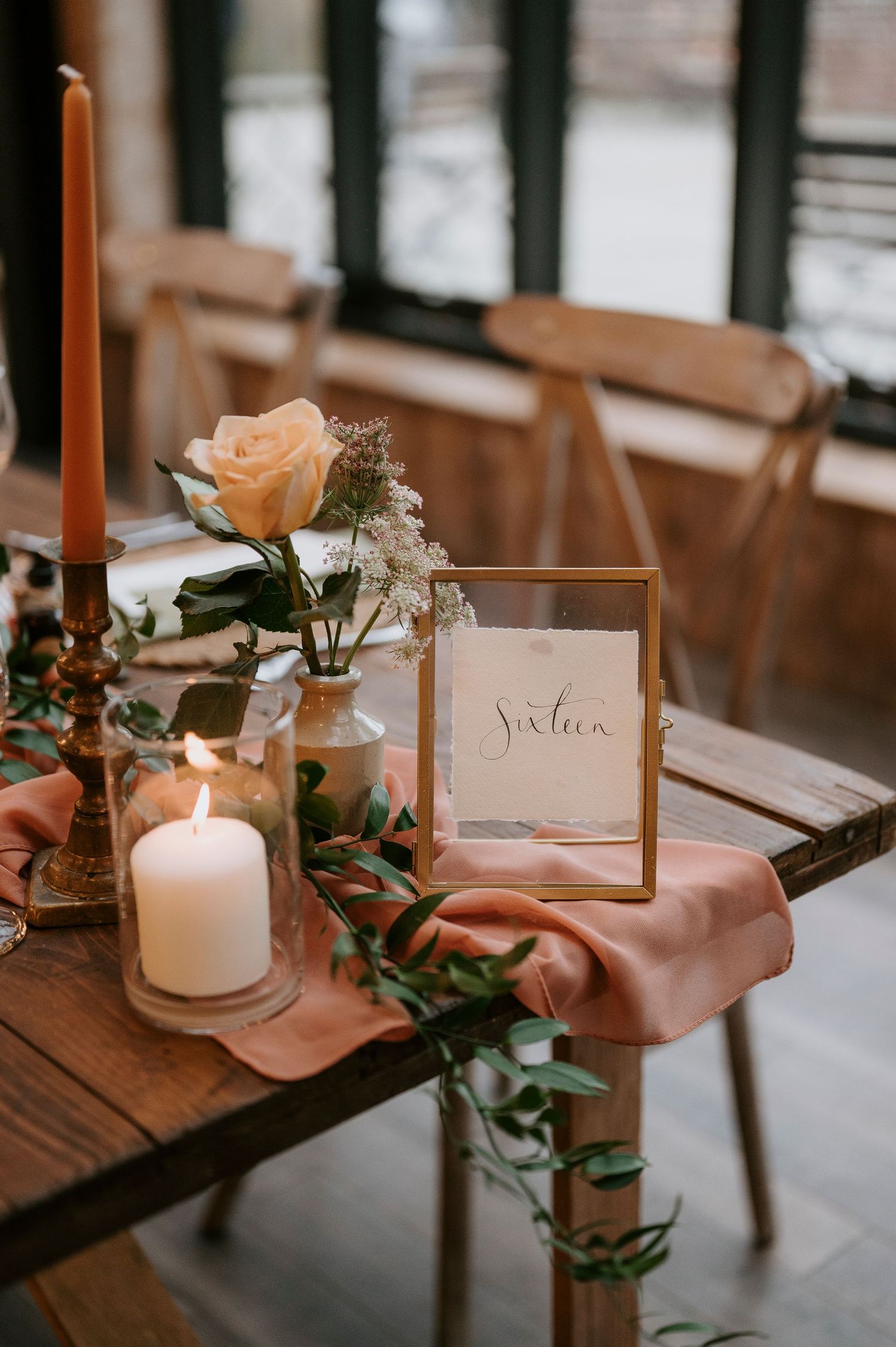 Elegant wedding table setting with peach linens, white candles, greenery and place cards on rustic wooden table.