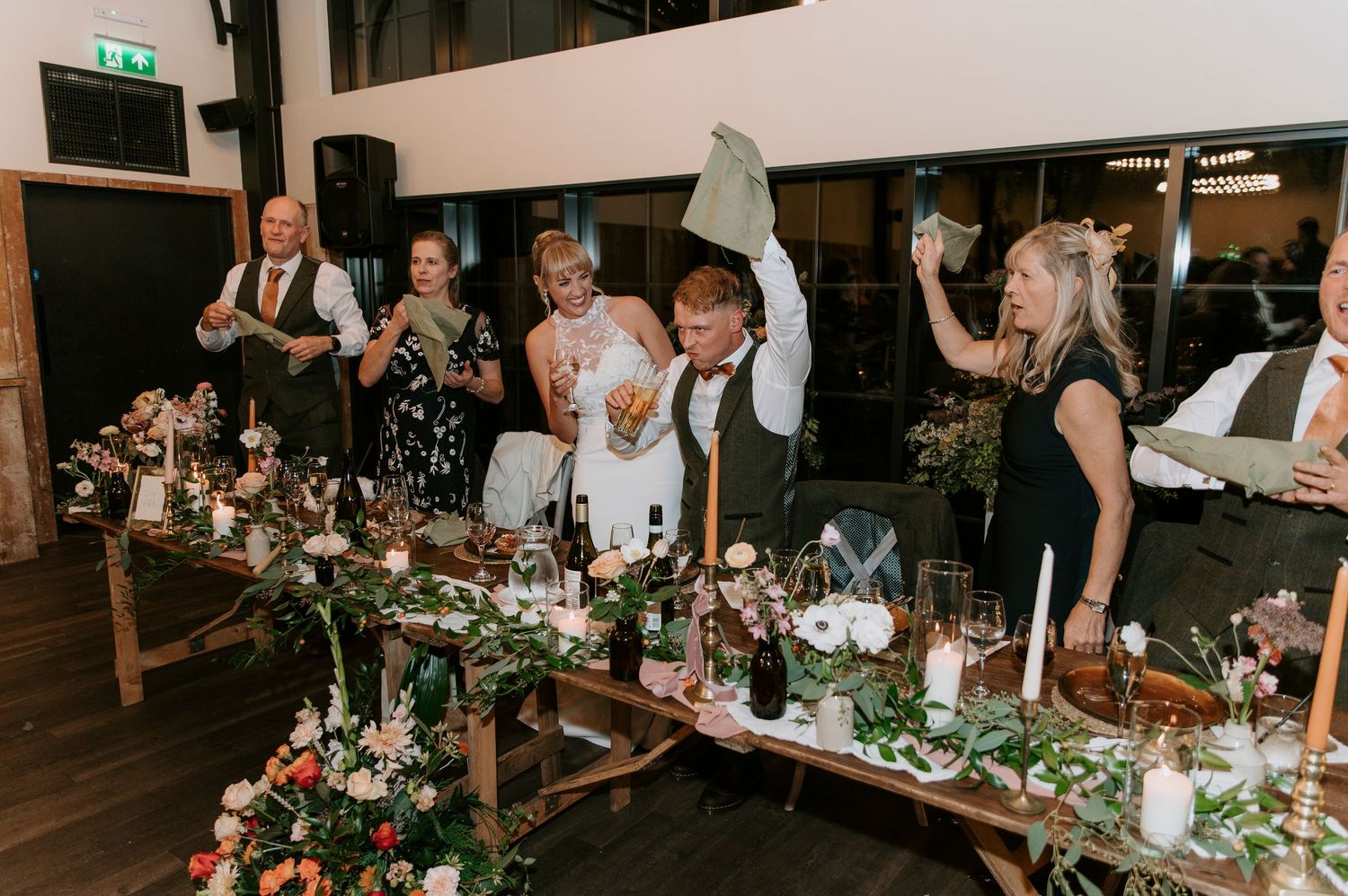 Wedding guests celebrate with raised glasses at a rustic reception table decorated with lush greenery and candlelight.