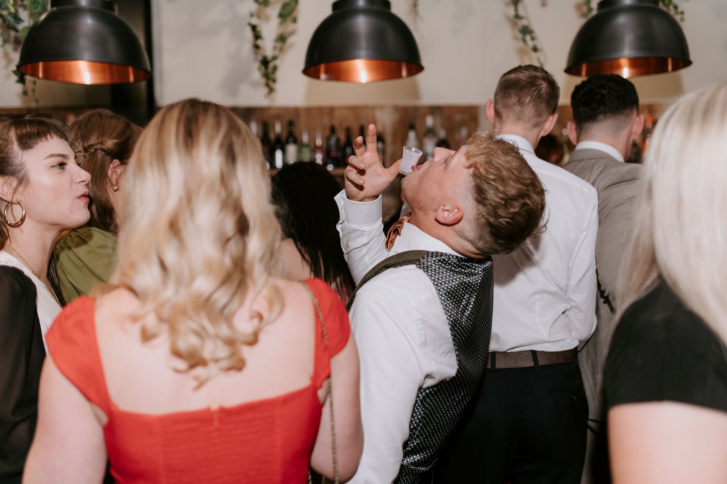 Wedding guests dance and celebrate at a reception under rustic pendant lighting and greenery decorations.
