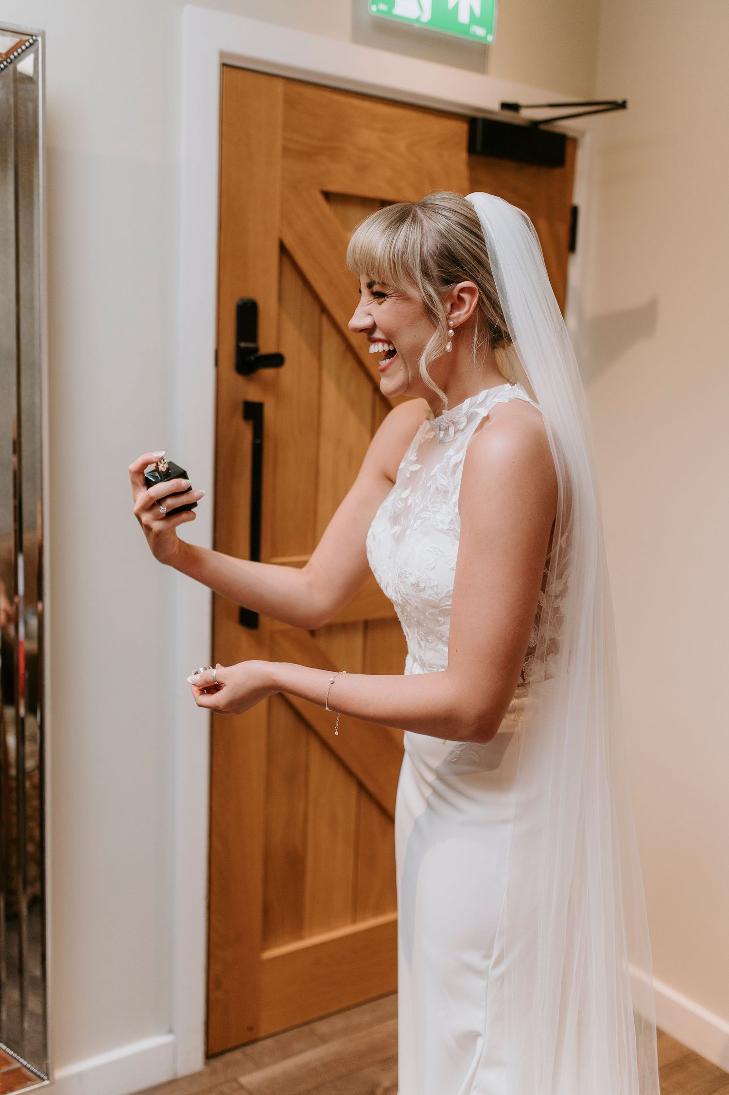 Bride in white lace dress checks her phone while standing in a hallway with wooden doors.