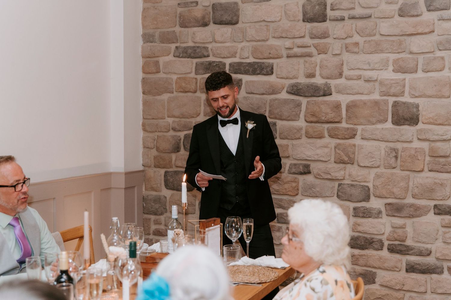 Wedding guests enjoy a reception dinner at long tables against a rustic stone wall backdrop.