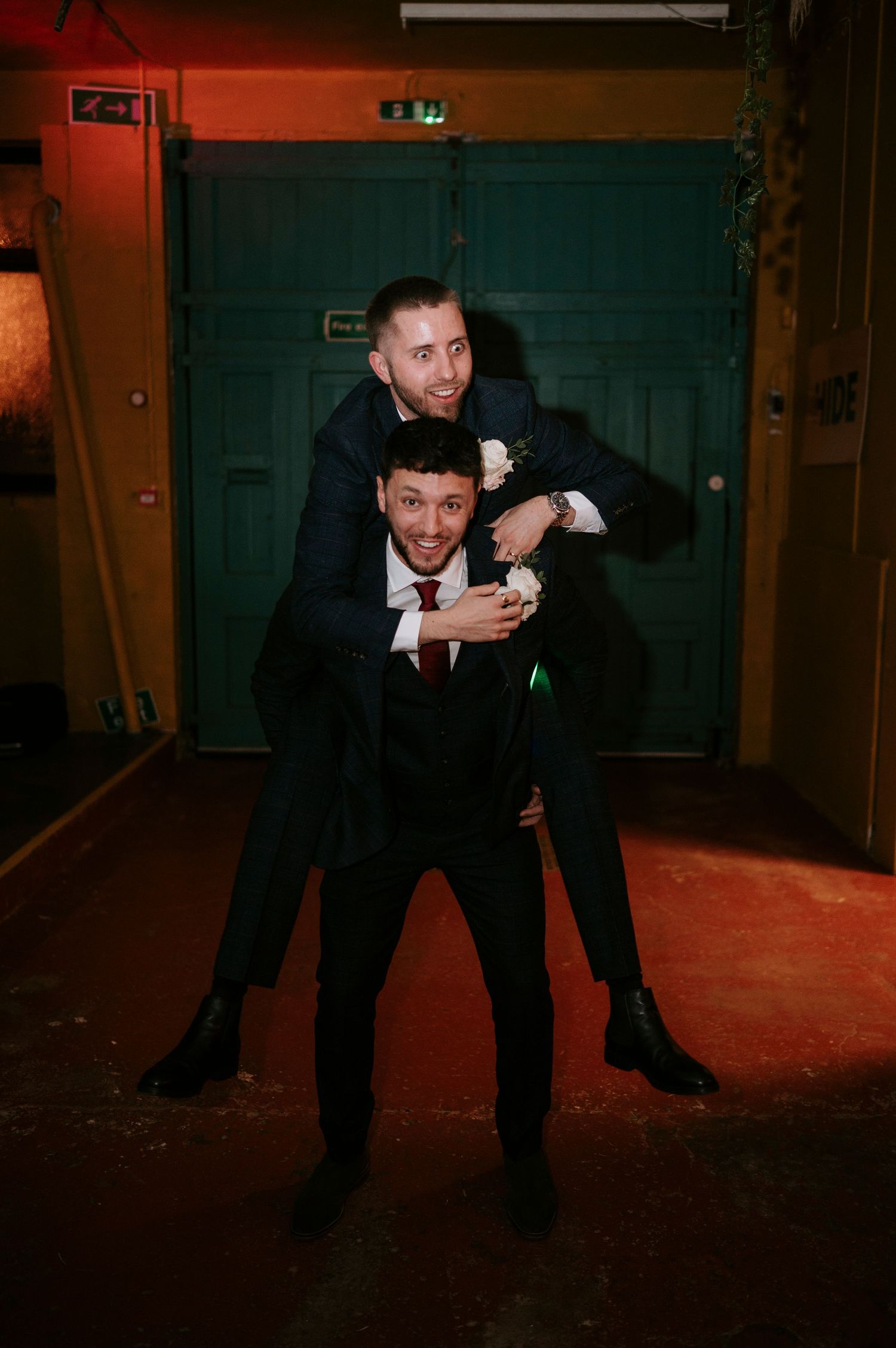 Two groomsmen in dark suits laughing as one gives the other a piggyback ride at wedding reception.