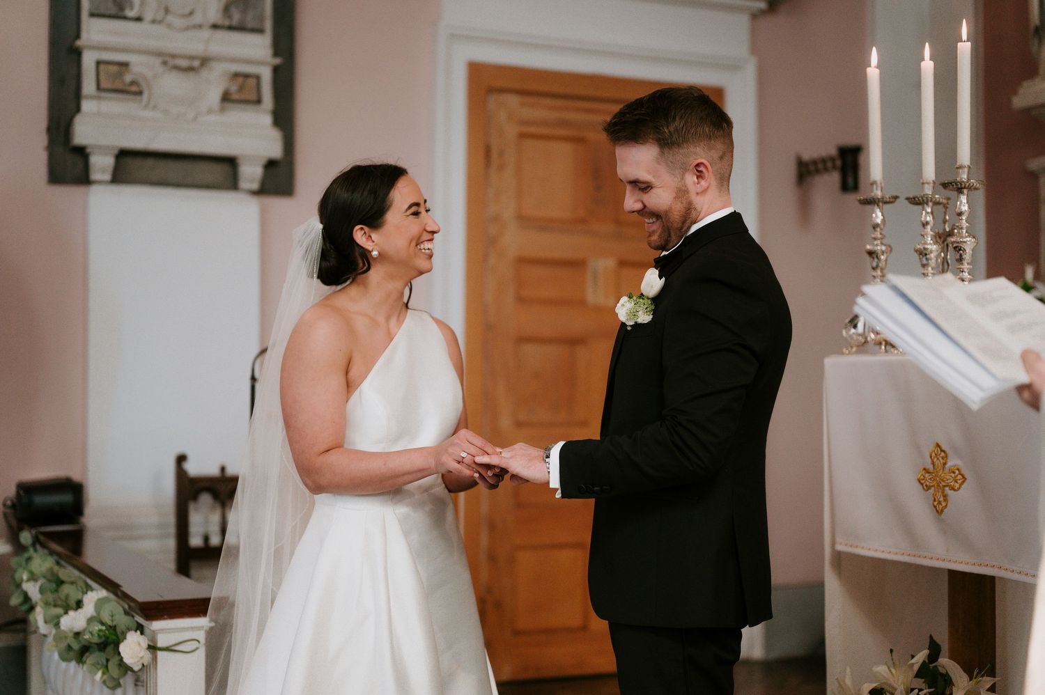 A couple exchanges vows during an intimate indoor wedding ceremony with white candles and floral decor.