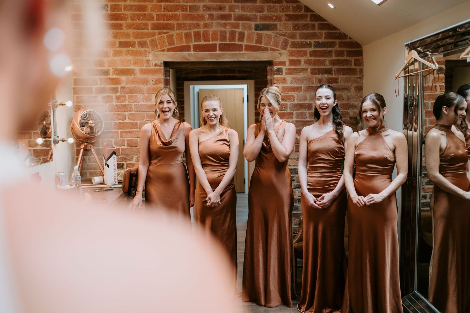 A row of bridesmaids in matching rust-colored dresses stand together in a brick-walled venue.