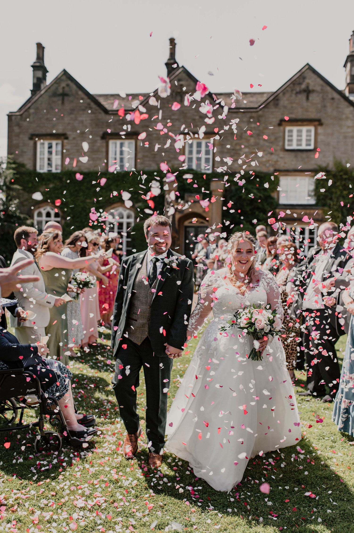 Wedding guests shower the newlyweds with pink rose petals on a sunny day outside a stone manor house.