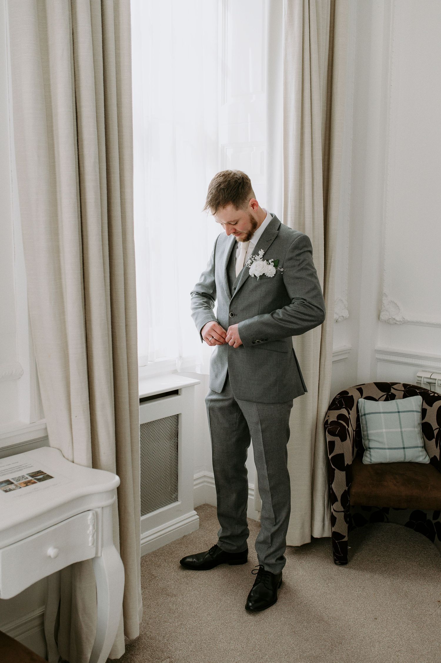 A person in a gray suit adjusts their tie while getting ready in a light-colored room with curtains.