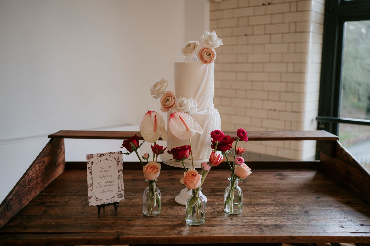 Small glass vases with pink and red roses line up on a rustic wooden table against white brick wall.