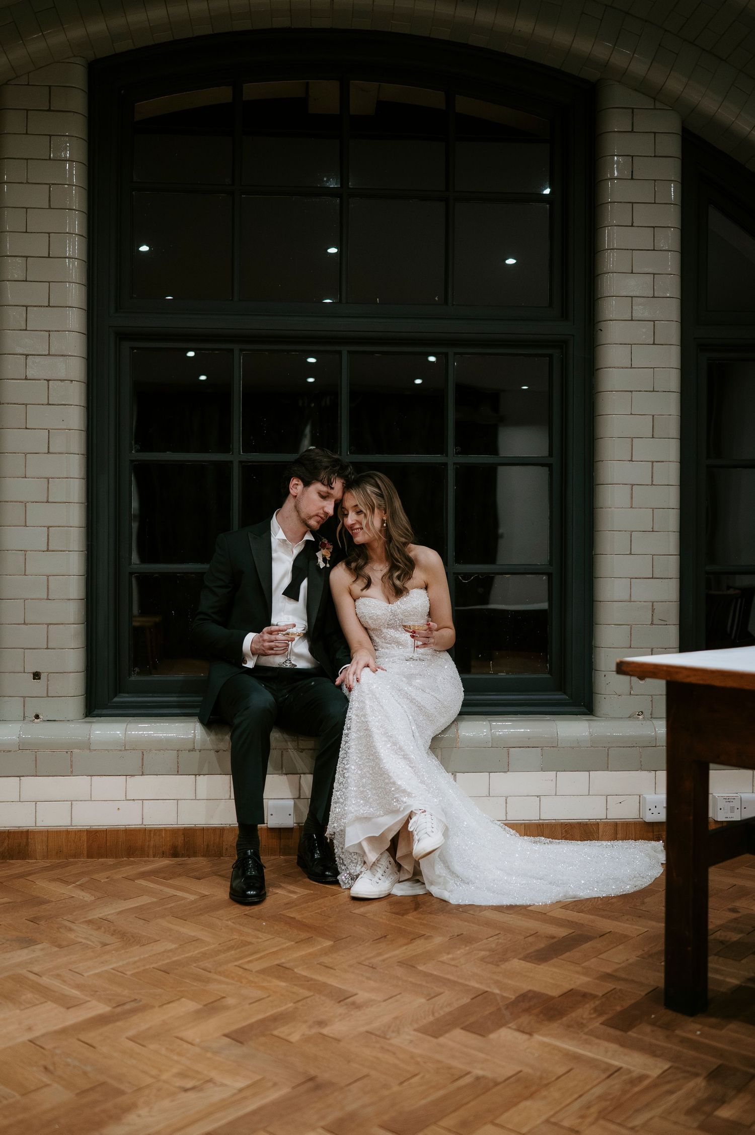 A couple shares an intimate moment while sitting together on a bench against white brick walls and herringbone wood floors.