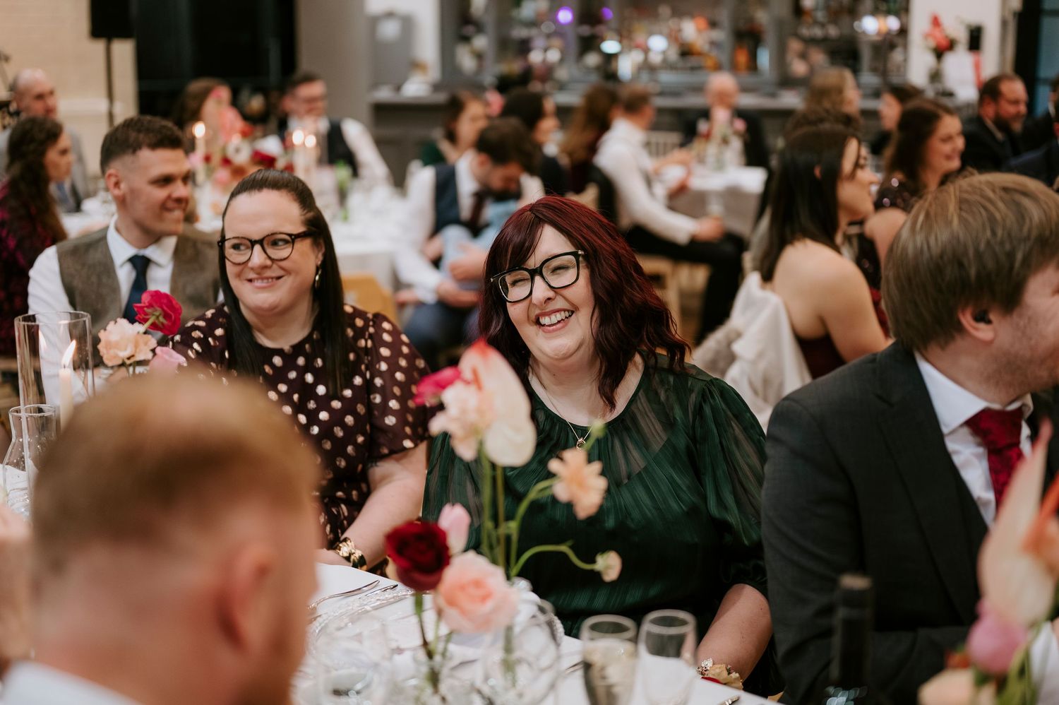 Wedding reception guests laugh and smile during dinner at elegantly decorated tables.