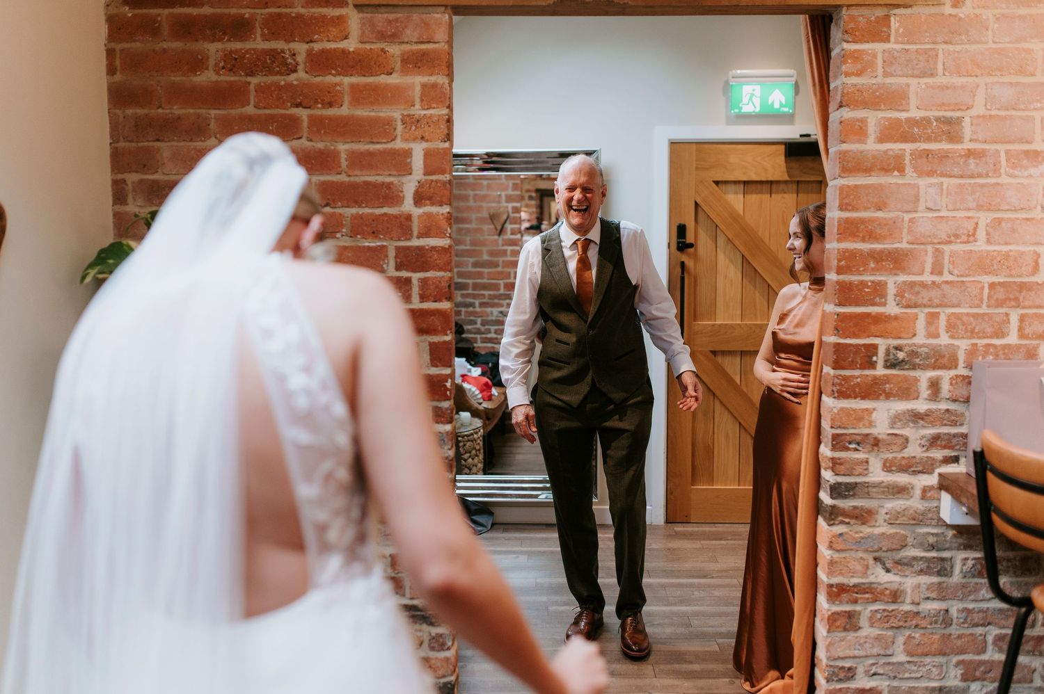 A bride in a white wedding dress stands in a rustic brick-walled venue as a person in a vest approaches.