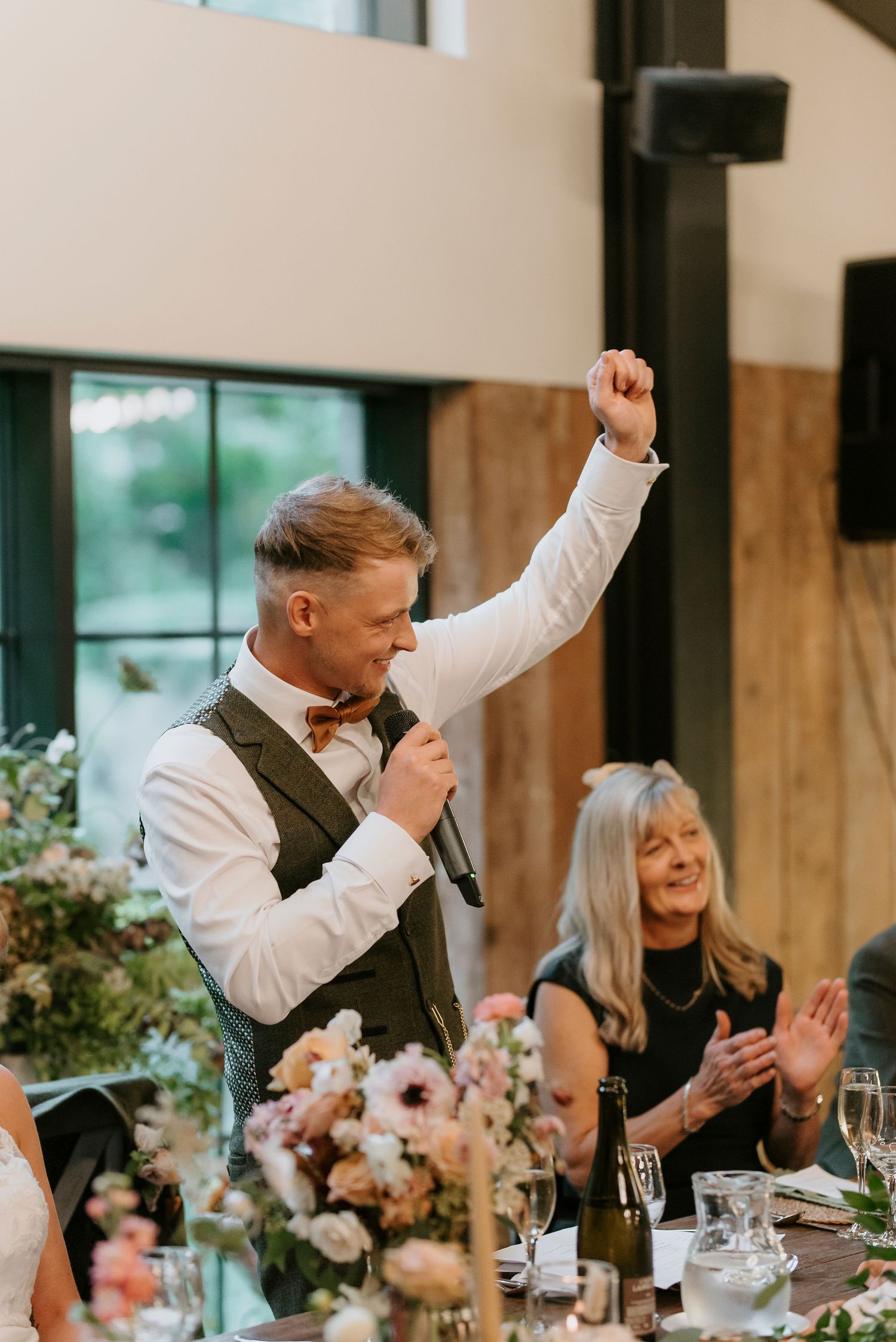 Wedding reception guests celebrate and cheer during emotional toasts at an elegant venue with floral centerpieces.