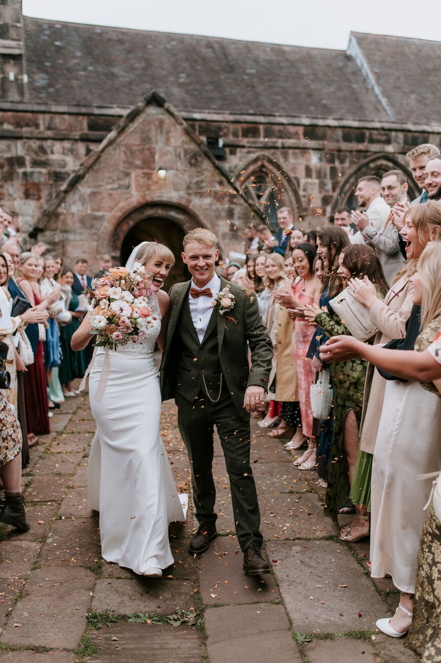 Newlyweds walk down a stone path outside a historic church while guests line both sides clapping and celebrating.