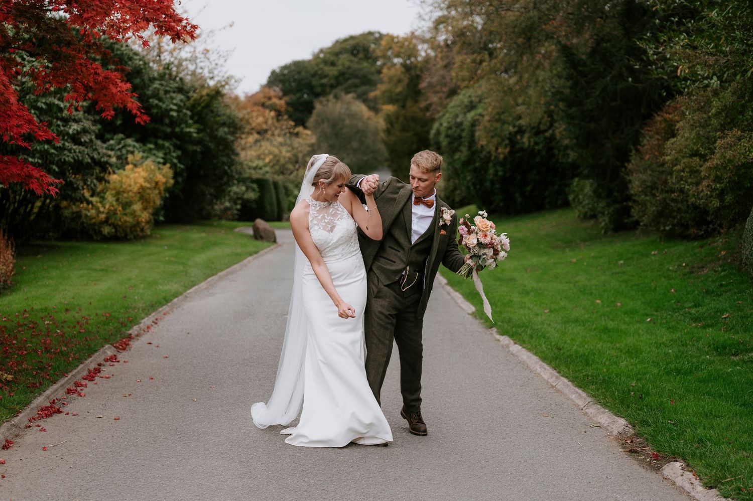 A couple in wedding attire walks along a tree-lined path with vibrant red autumn foliage overhead.
