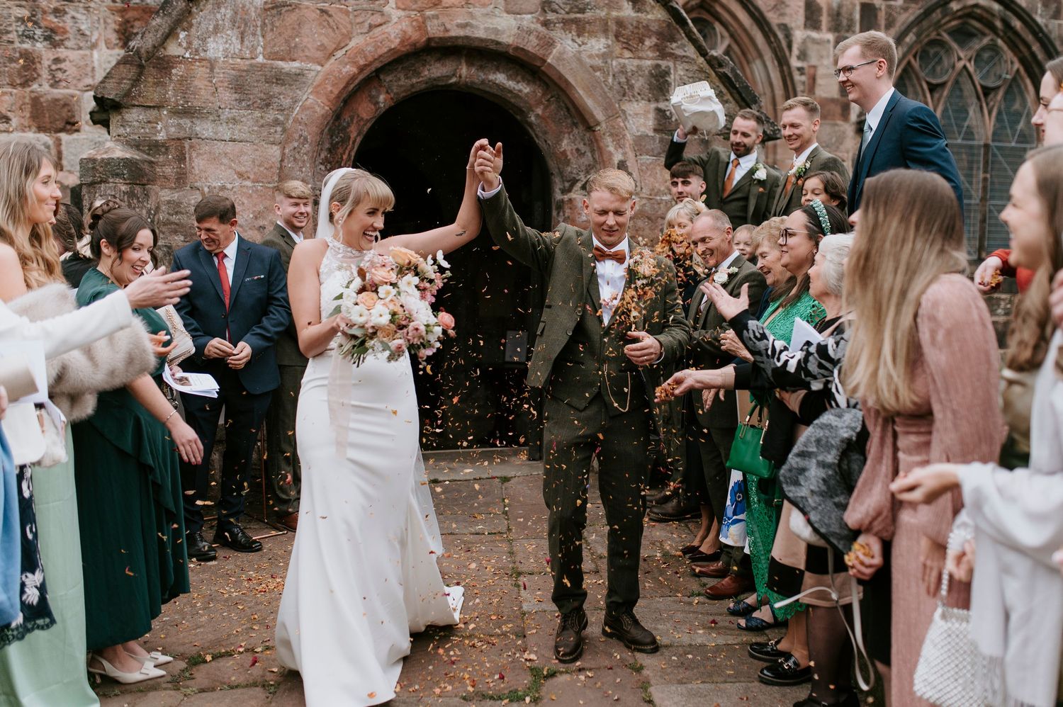 Wedding guests throw flower petals in celebration as newlyweds exit stone church on their special day.