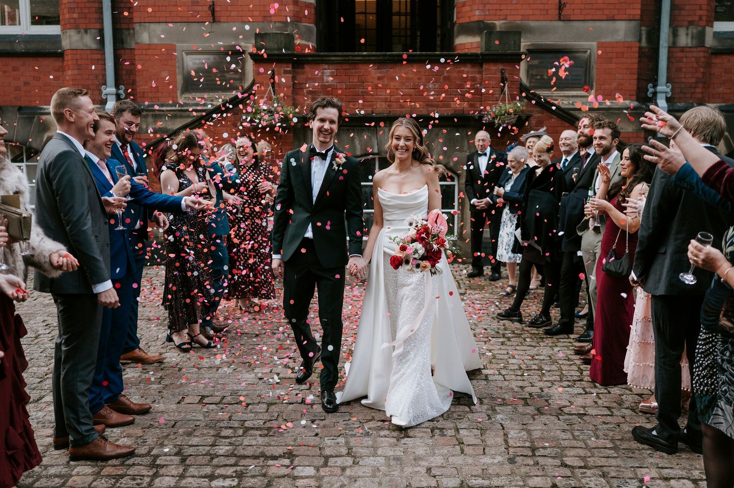 Wedding guests shower newlyweds with red rose petals on cobblestone street outside brick building.