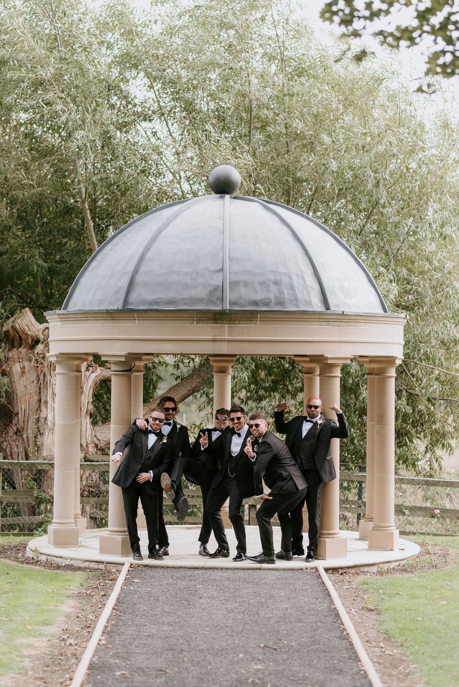 A wedding party poses playfully in a series of photos under an elegant domed gazebo in a garden setting.