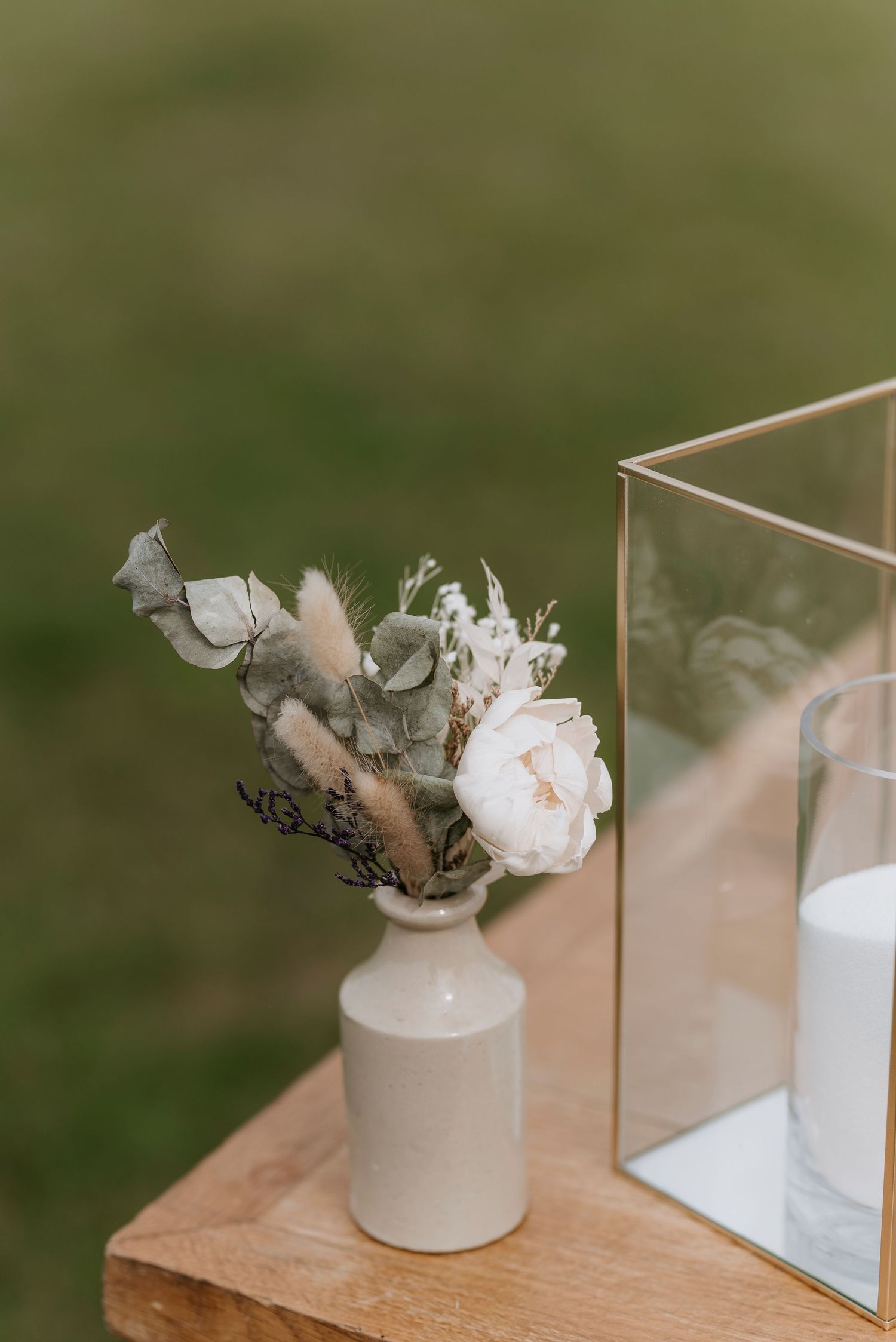 A delicate white flower arrangement in a small ceramic vase sits beside a glass container on a beige surface.