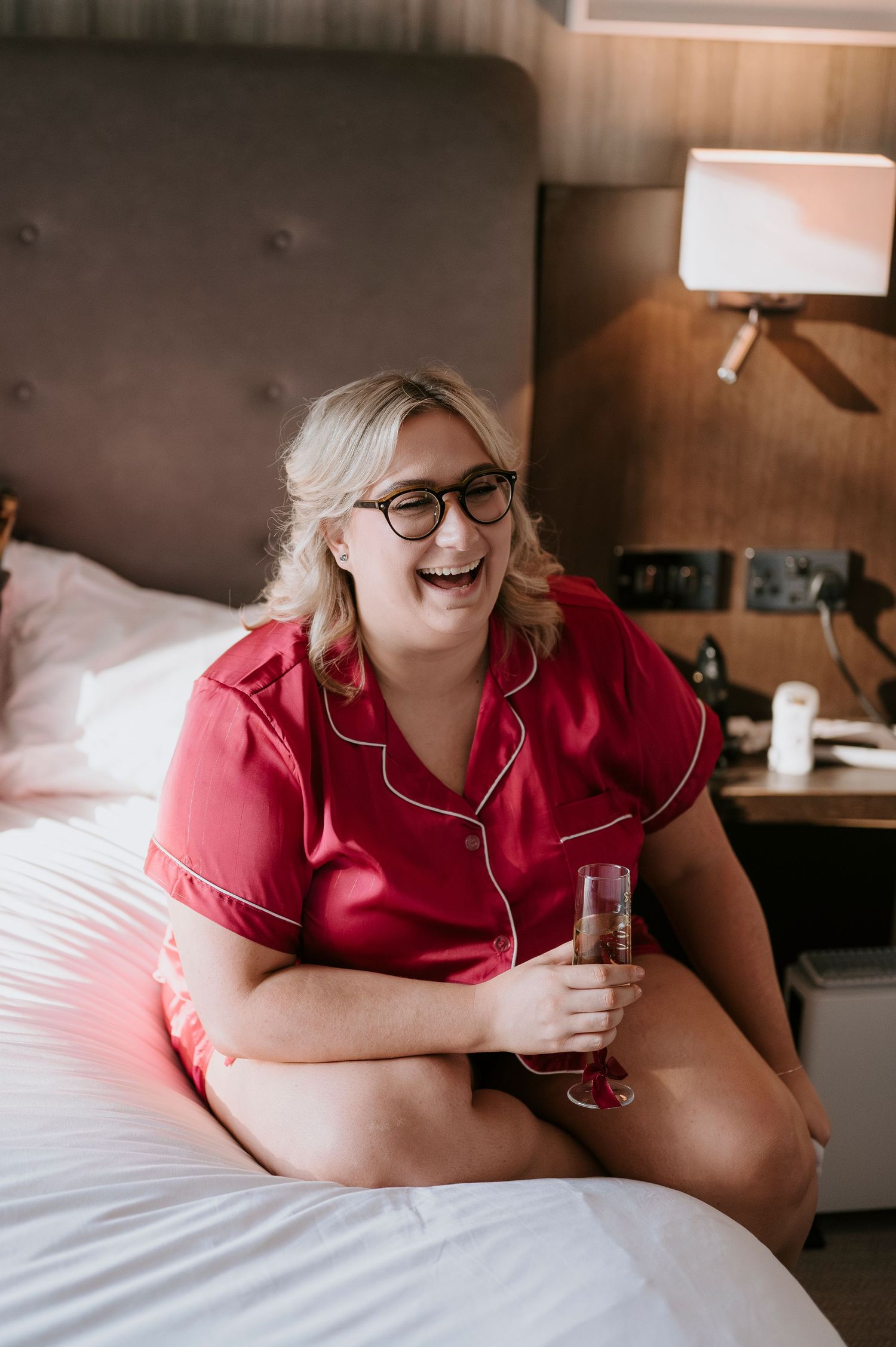 A laughing blonde woman in red satin pajamas sits on a hotel bed holding a champagne flute.
