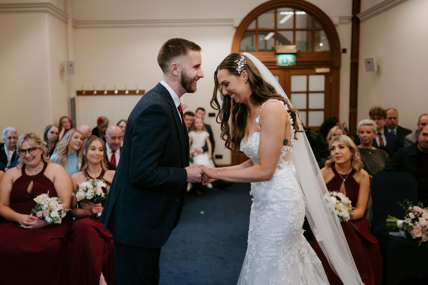 A bride and groom laugh joyfully while exchanging vows at their wedding ceremony, with bridesmaids in burgundy dresses behind them.