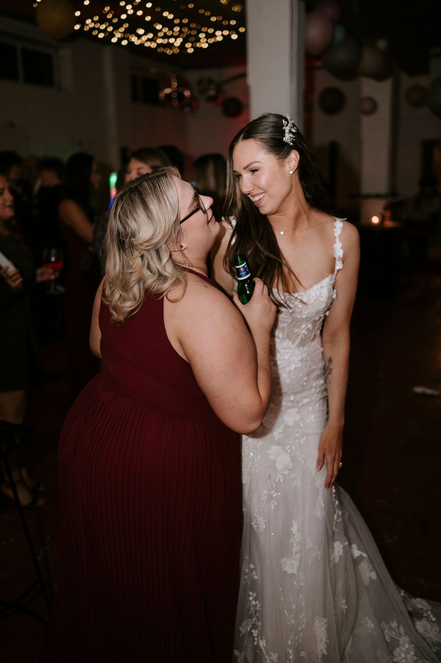 A bride in a lace gown shares a joyful moment with a guest in burgundy at a wedding reception.