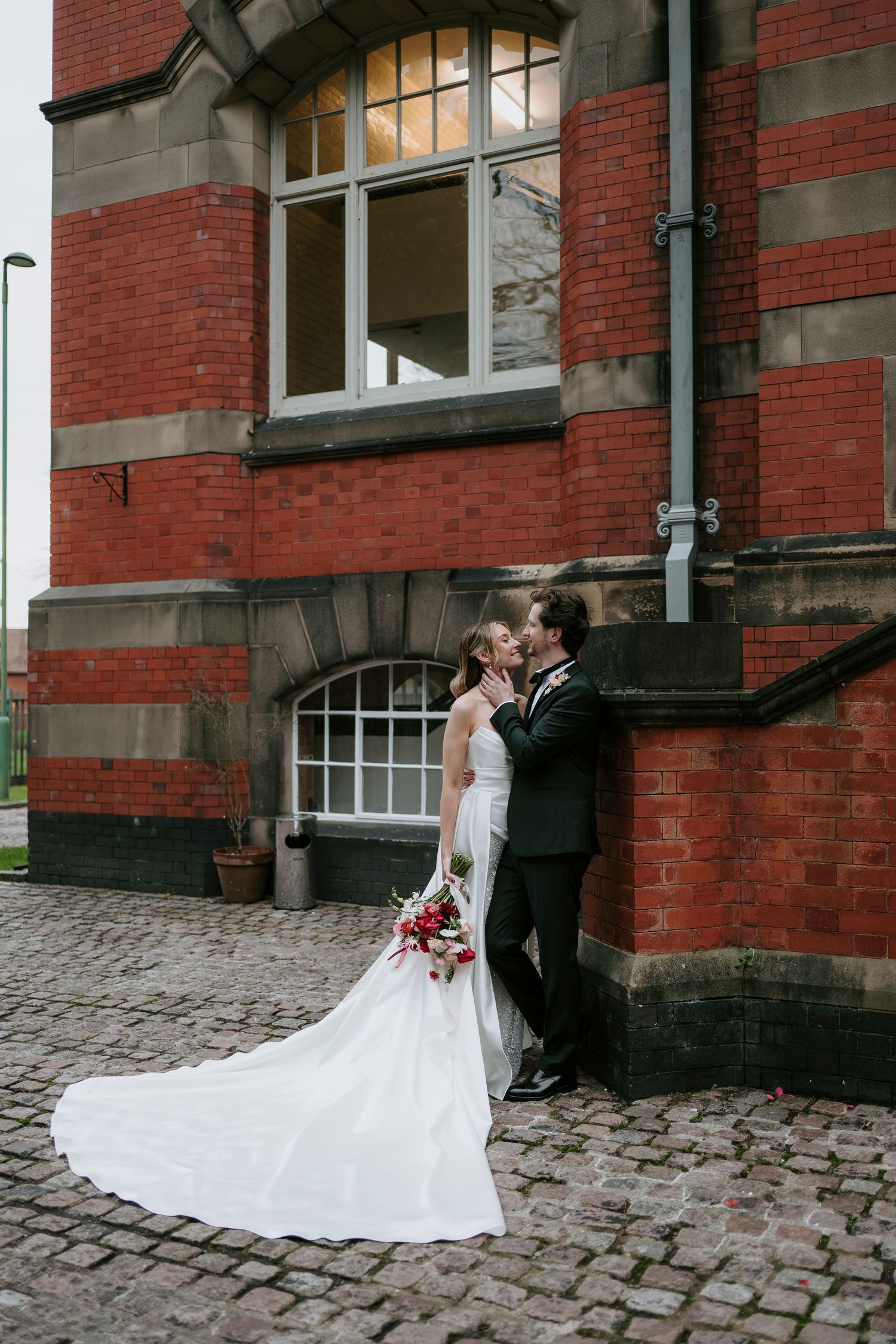 A couple in wedding attire shares a romantic moment against a red brick building with cobblestone ground.
