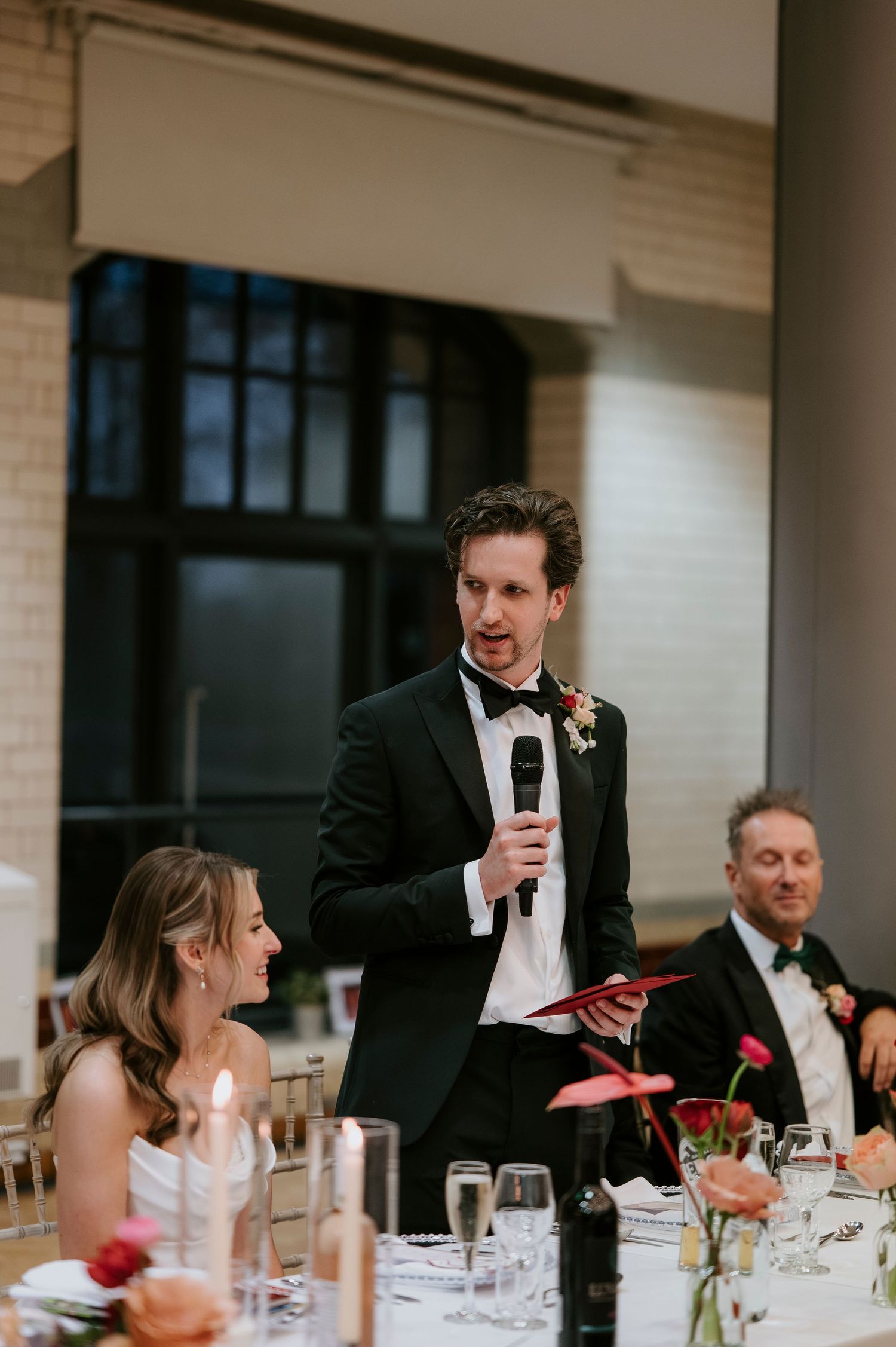 Wedding reception guests seated at elegant tables with red floral centerpieces listen to speeches during dinner.