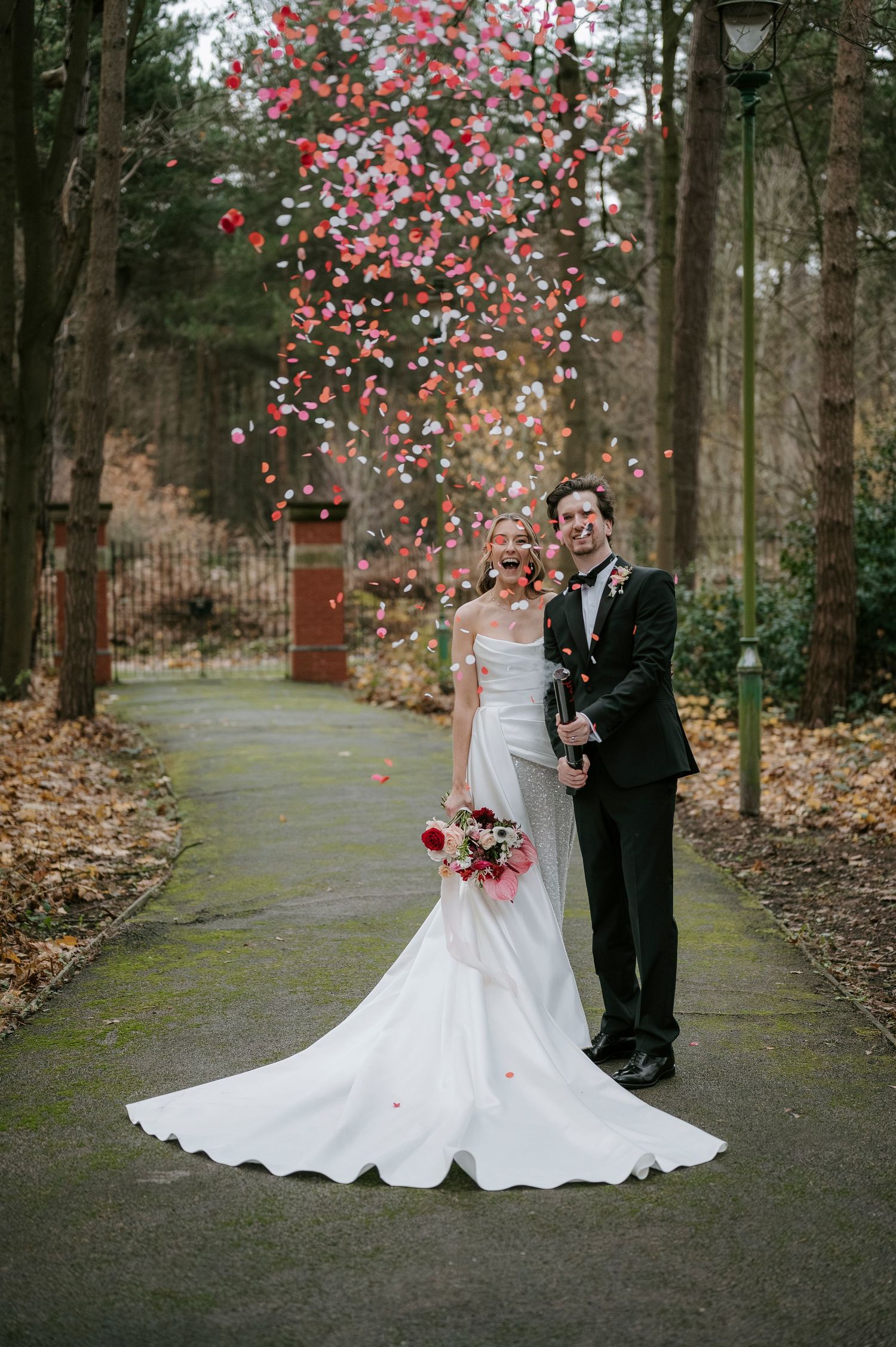 Red rose petals shower over newlyweds on a forest path during their romantic photo session.