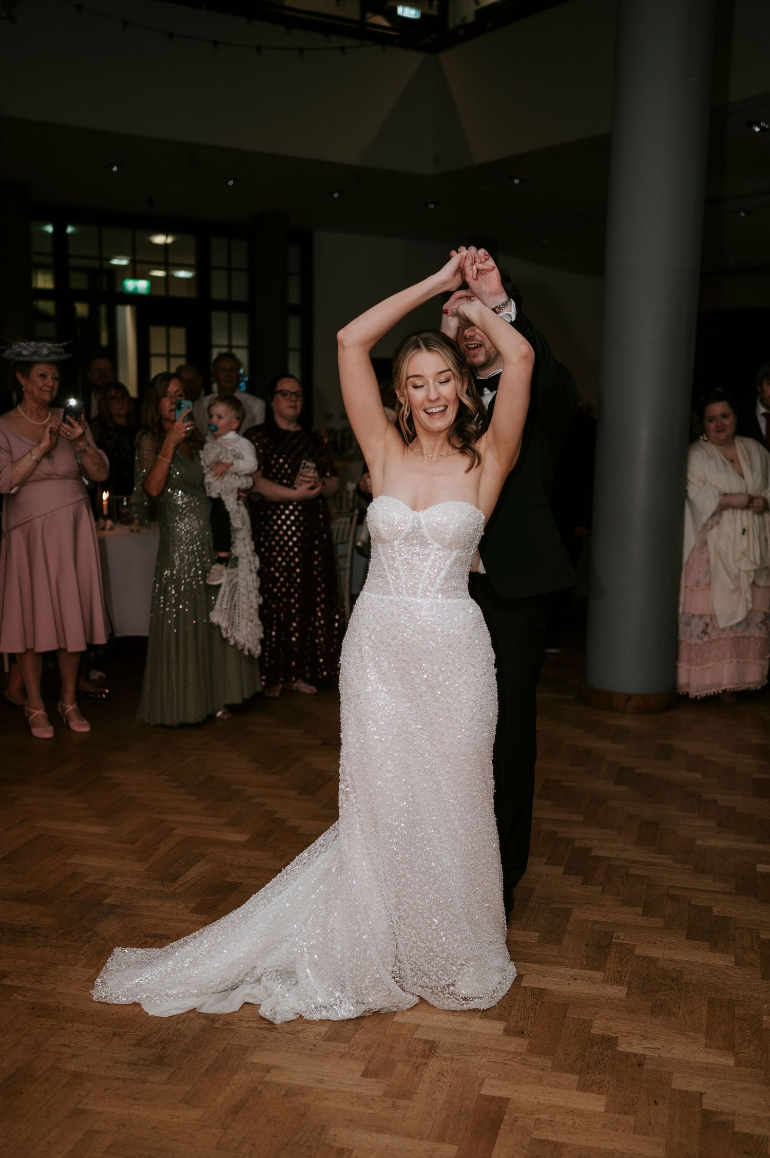 A bride in a sparkly white wedding dress dances joyfully on a wooden dance floor during a wedding reception.
