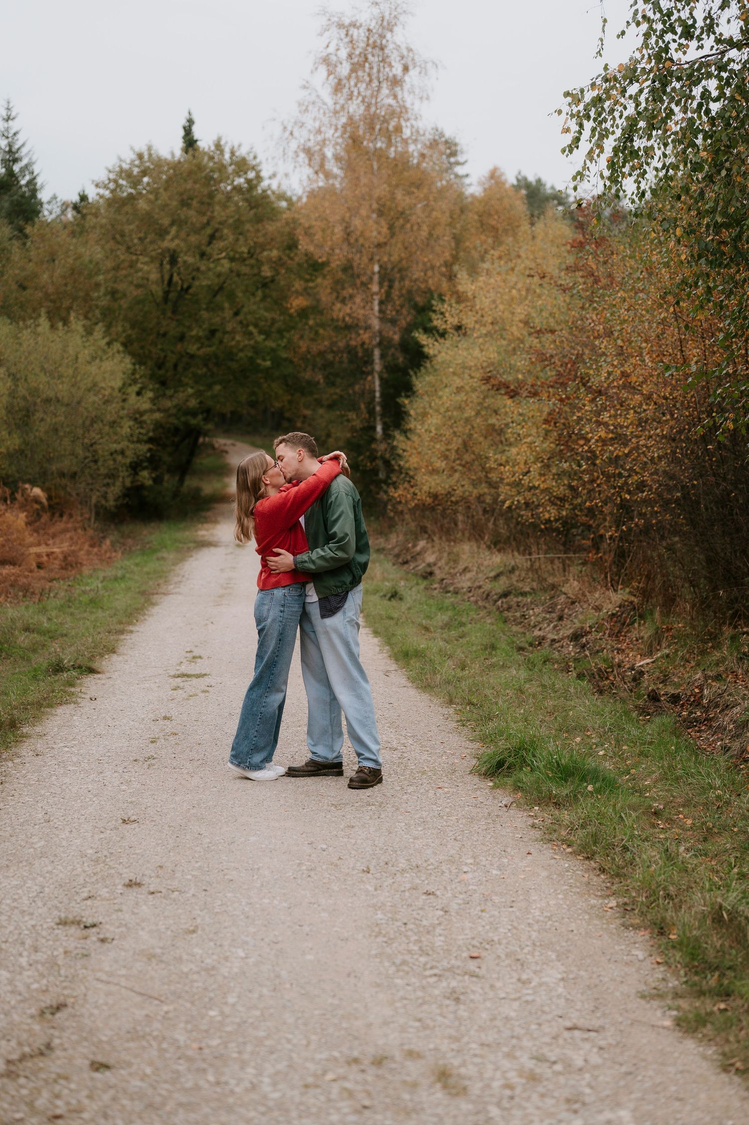 A couple embraces on a dirt path surrounded by autumn trees with golden foliage on a cloudy day.