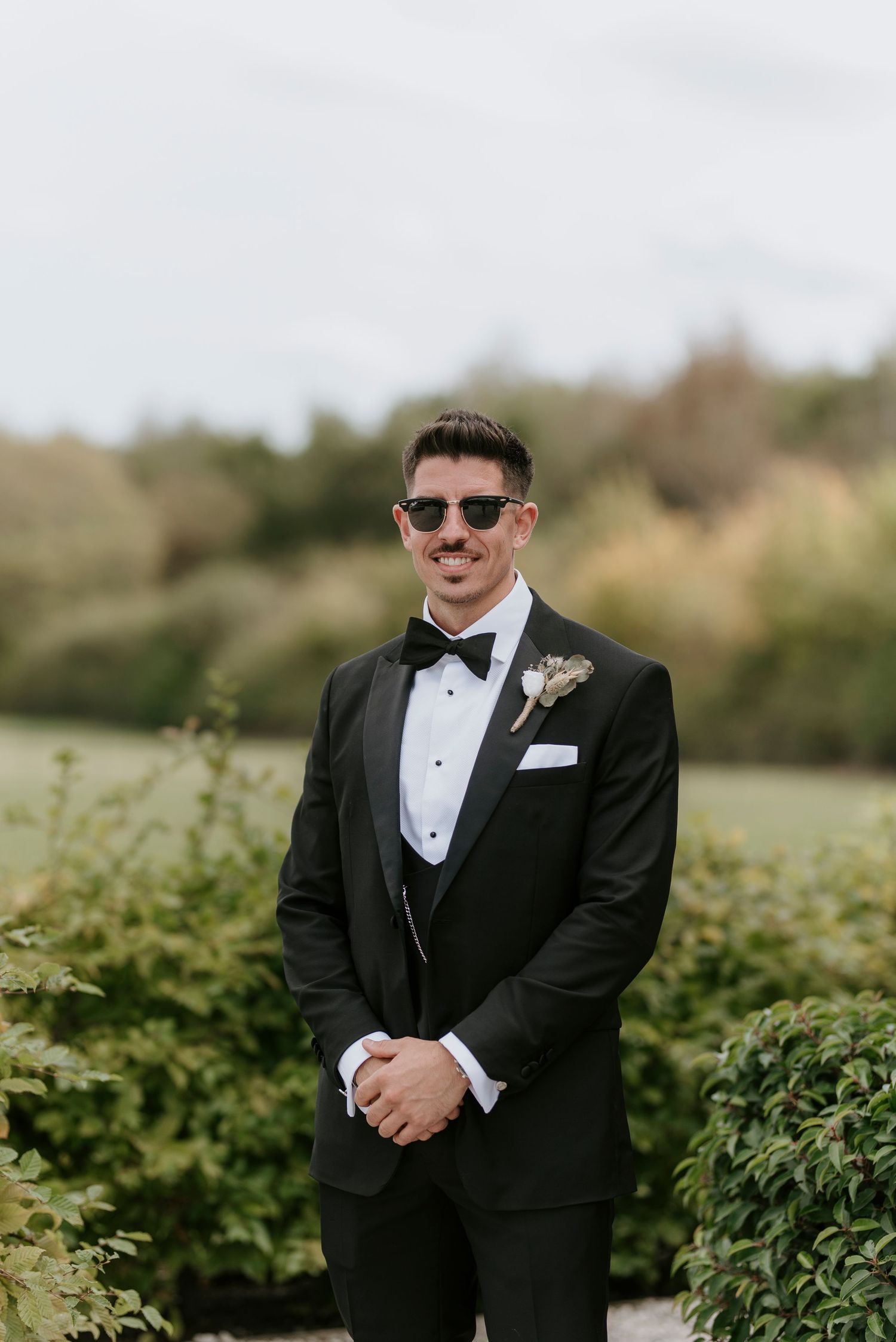 A groom in a black tuxedo and sunglasses poses outdoors in front of green foliage on his wedding day.