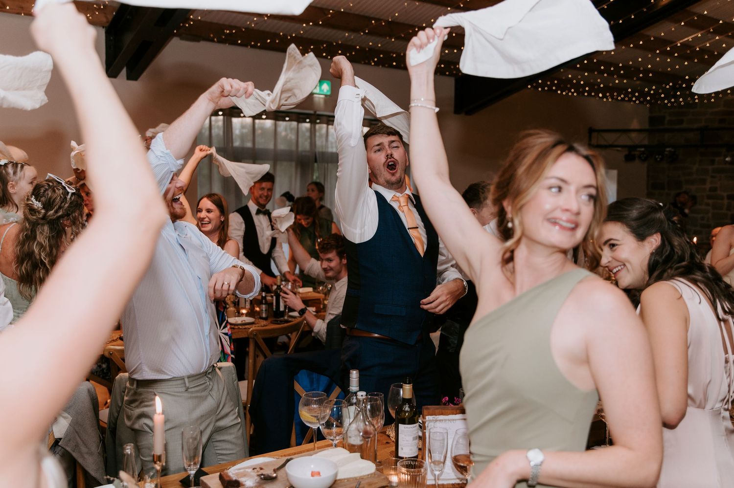Wedding guests dance and celebrate with raised arms on a crowded dance floor at an indoor reception venue.