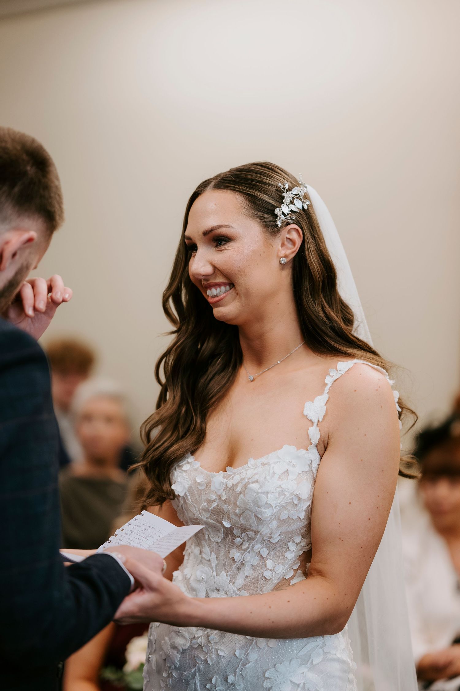 Smiling bride in floral lace wedding dress and veil reads vows during indoor wedding ceremony.