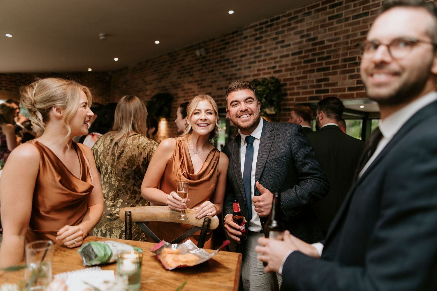 People laughing and socializing at a wedding reception around wooden tables in a rustic brick-walled venue.