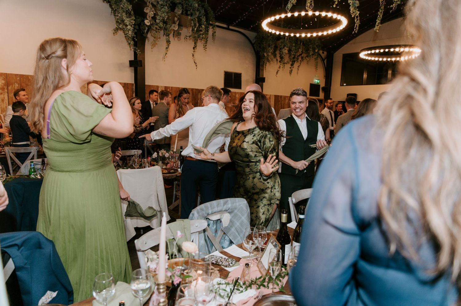 Wedding reception guests dance and celebrate under elegant circular chandeliers in a rustic venue space.