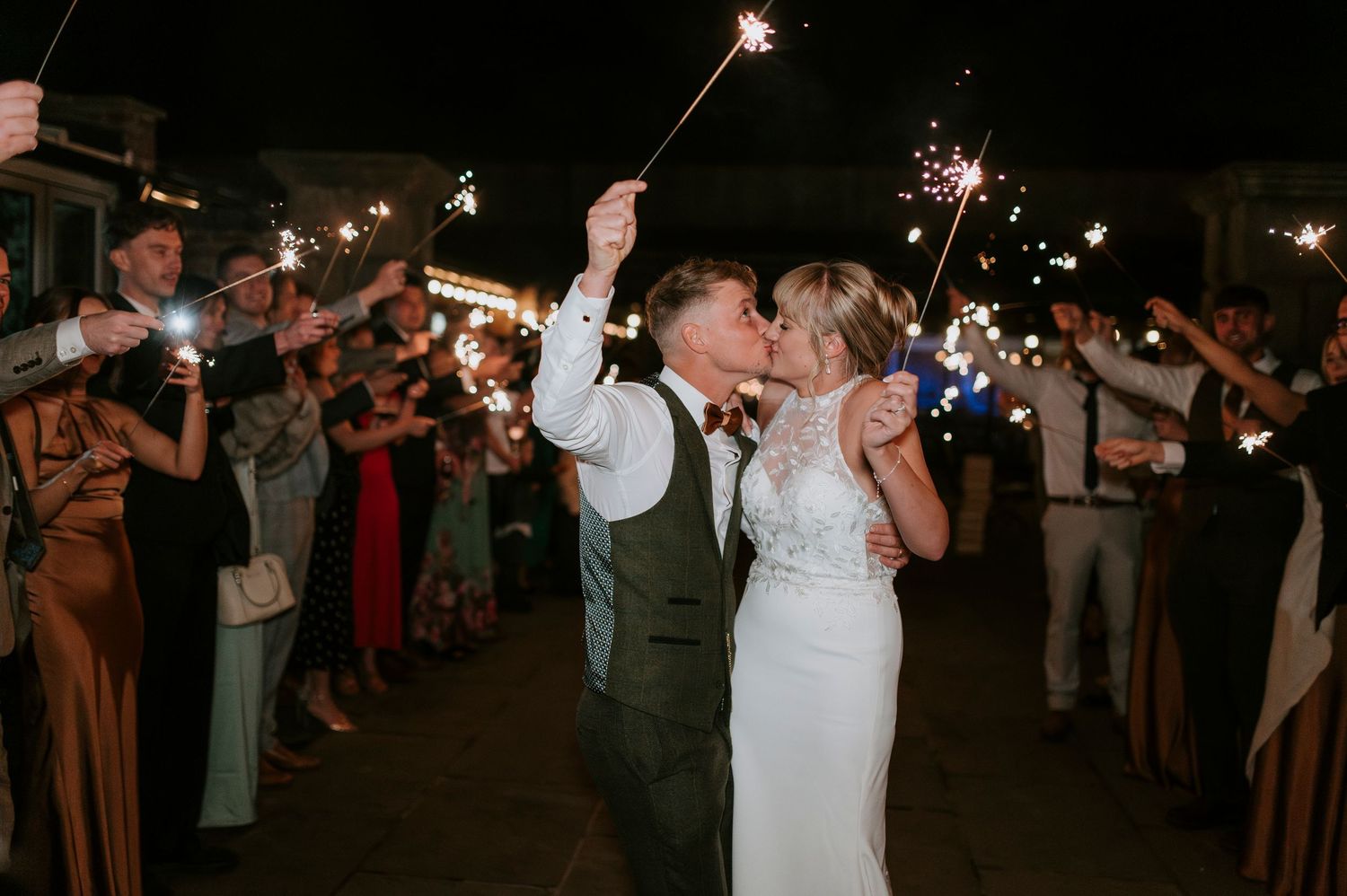Wedding guests hold sparklers creating a magical nighttime send-off tunnel for the newlyweds.