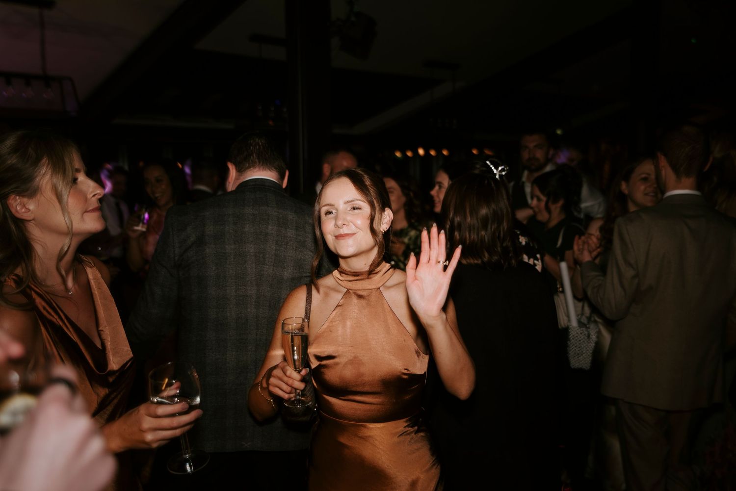 People dancing and socializing at a dimly lit party with someone in a brown satin dress waving to the camera.