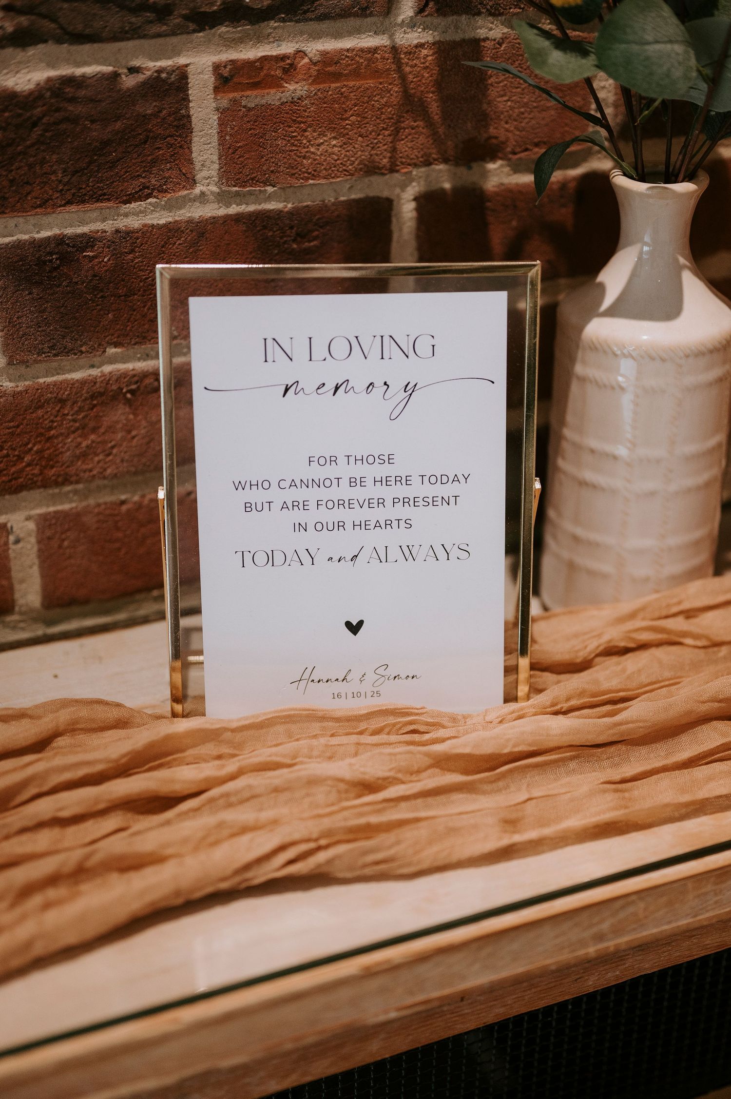 Memorial signs and white vases displayed on a wooden shelf against a brick wall at a wedding ceremony.