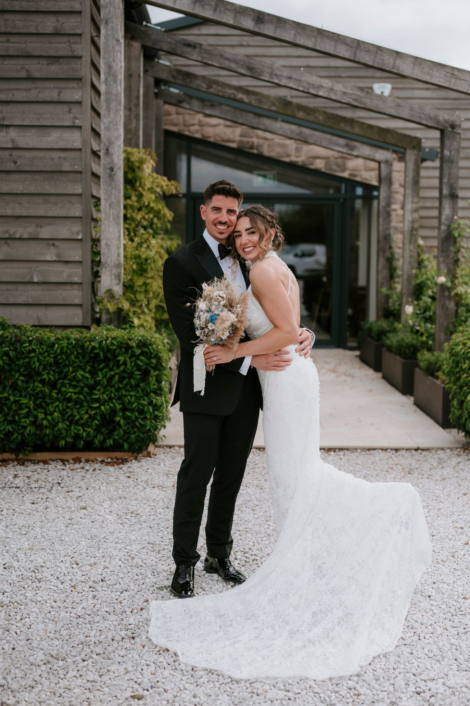 A couple shares an intimate moment in their wedding attire on a white gravel path by a rustic wooden building.